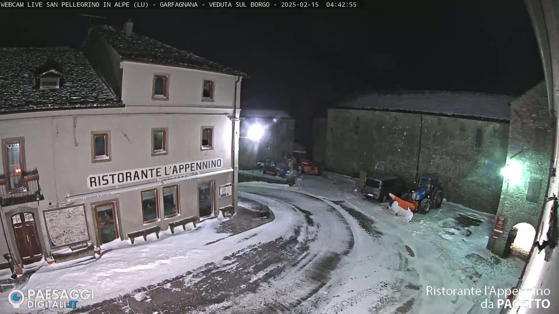 A snow-covered town square at night, illuminated by streetlights, with a restaurant and a snowplow parked nearby.