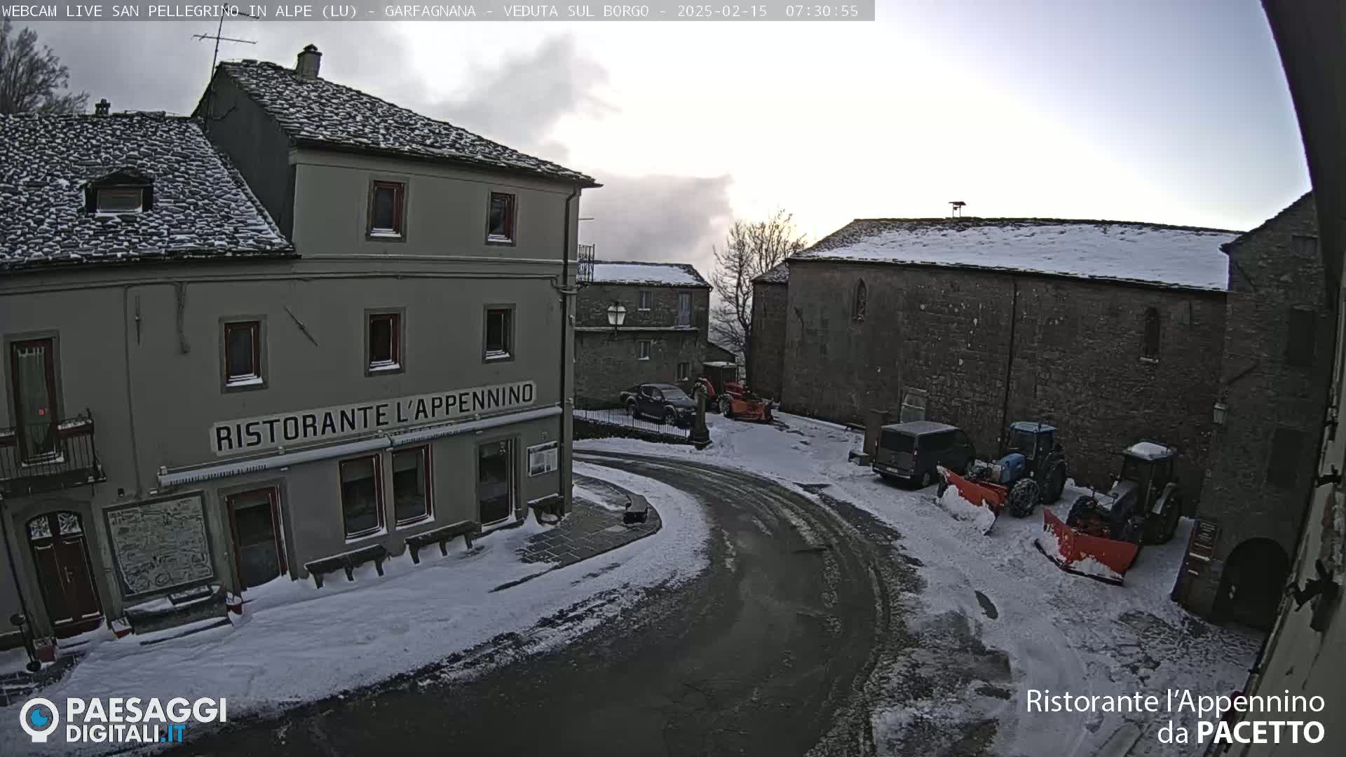 A snow-covered village square with several buildings, some vehicles, and snow-clearing equipment.