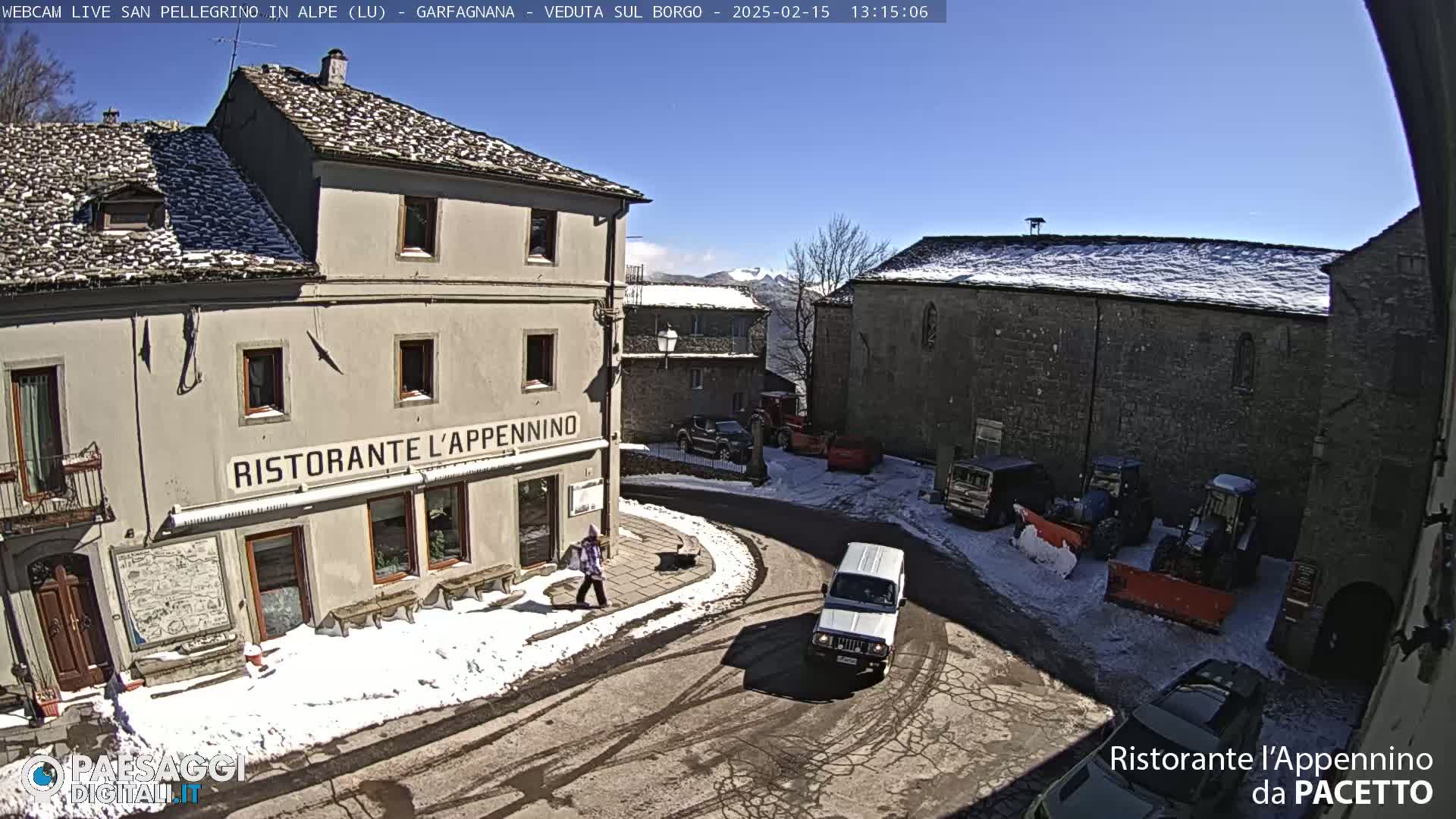 A sunny, snow-covered village square with several buildings, some vehicles, and a person walking.