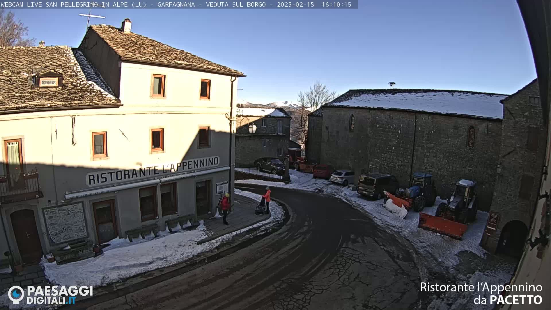 A snow-dusted village square with stone buildings, parked cars and tractors, and two people walking near a restaurant.
