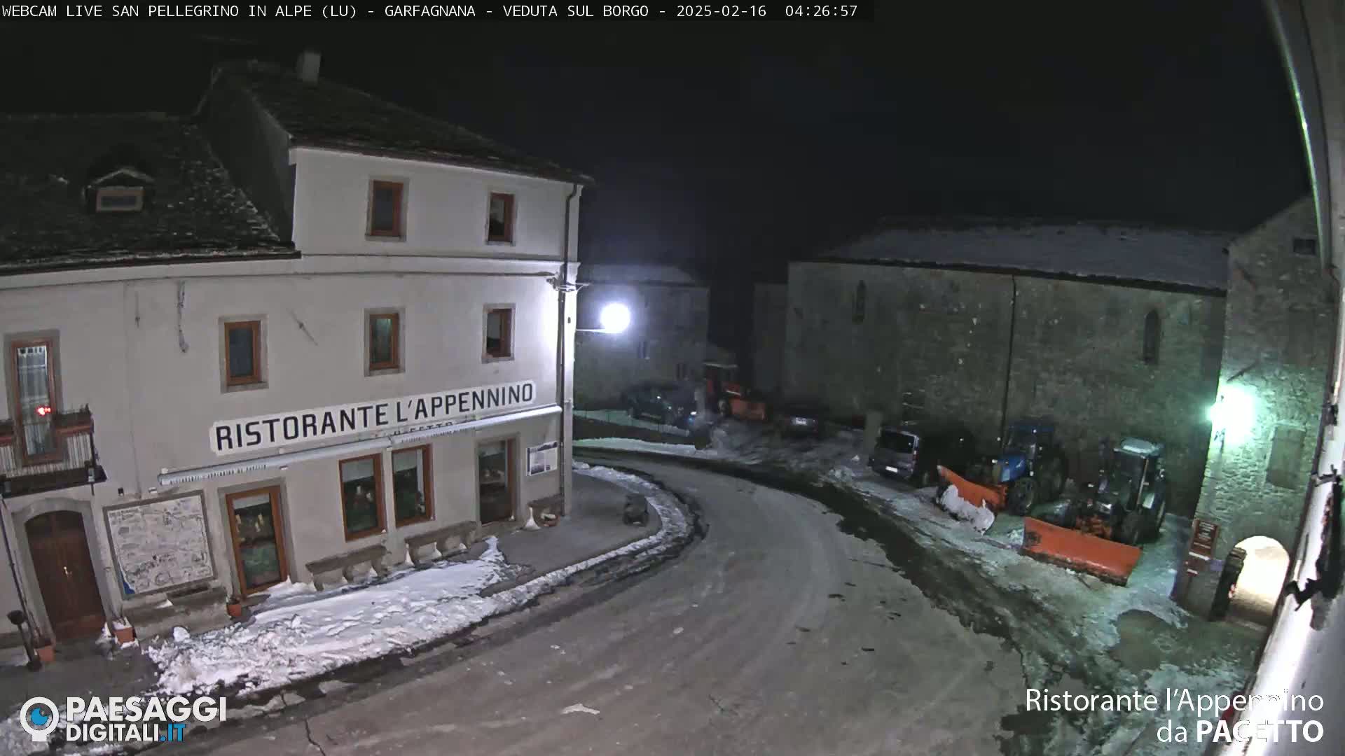 A snow-covered village square at night, with a restaurant and parked vehicles, is illuminated by streetlights.