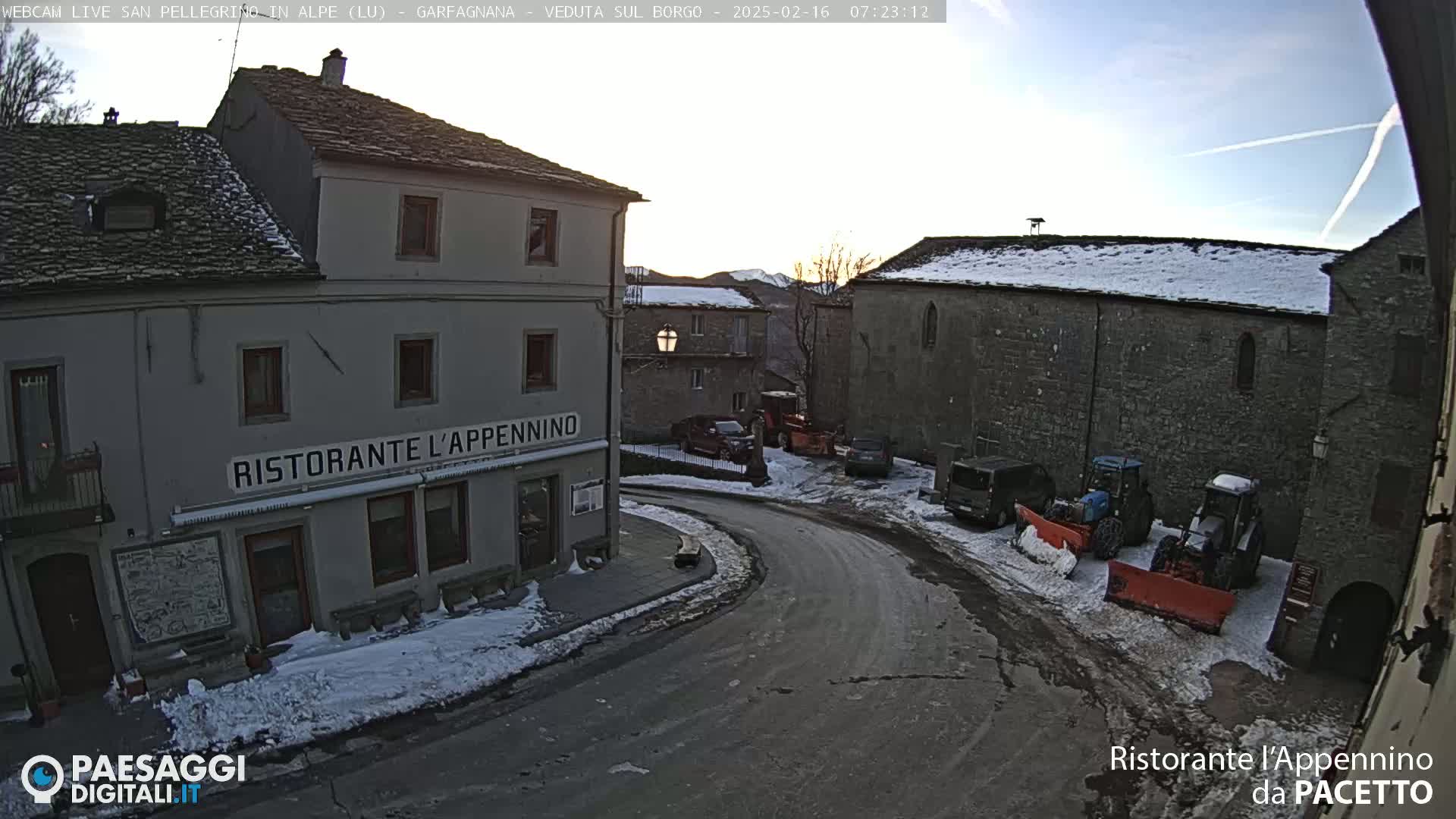 A snow-covered village square with several buildings, parked vehicles, and snow plows under a partly clear sky.