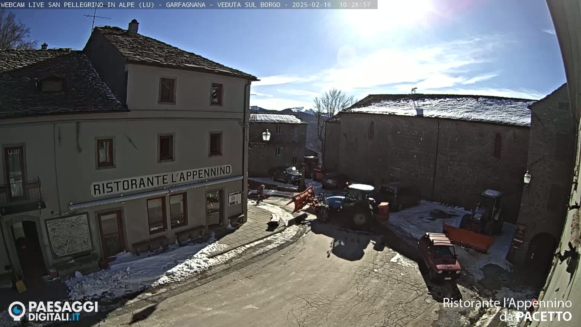 A sunny, snowy village square with a tractor and snow plow, several buildings including a restaurant, and a few parked cars.