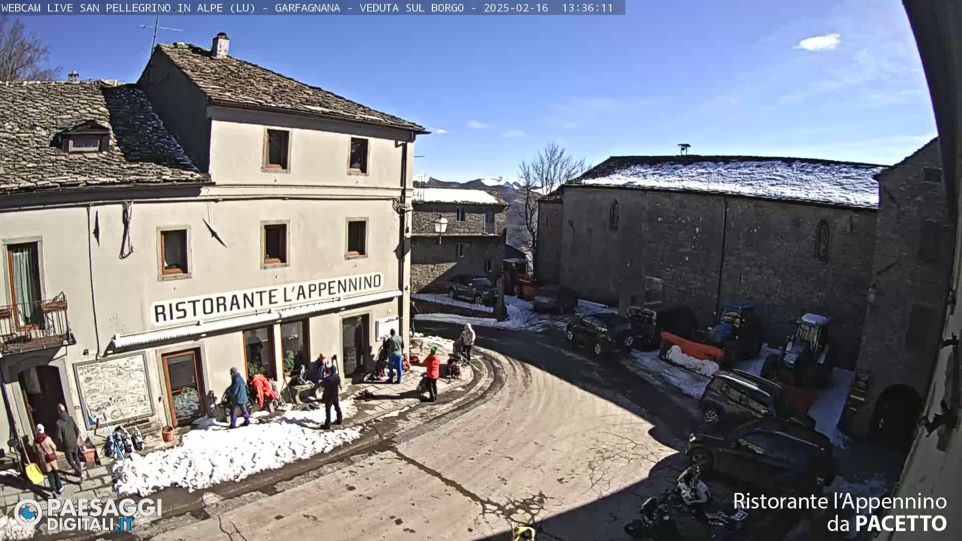 A sunny day in a snow-dusted village square with several people gathered near a restaurant, vehicles parked nearby, and farm equipment visible in the background.