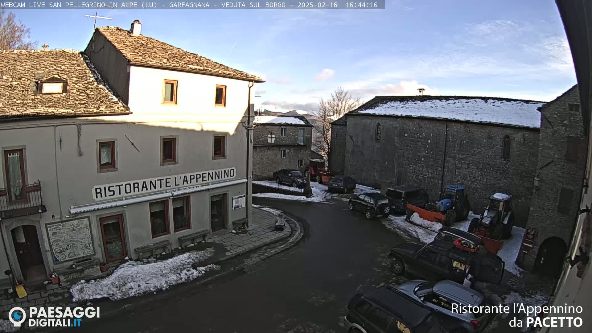 A snow-dusted village square with several parked cars and farm equipment, under a mostly clear sky.
