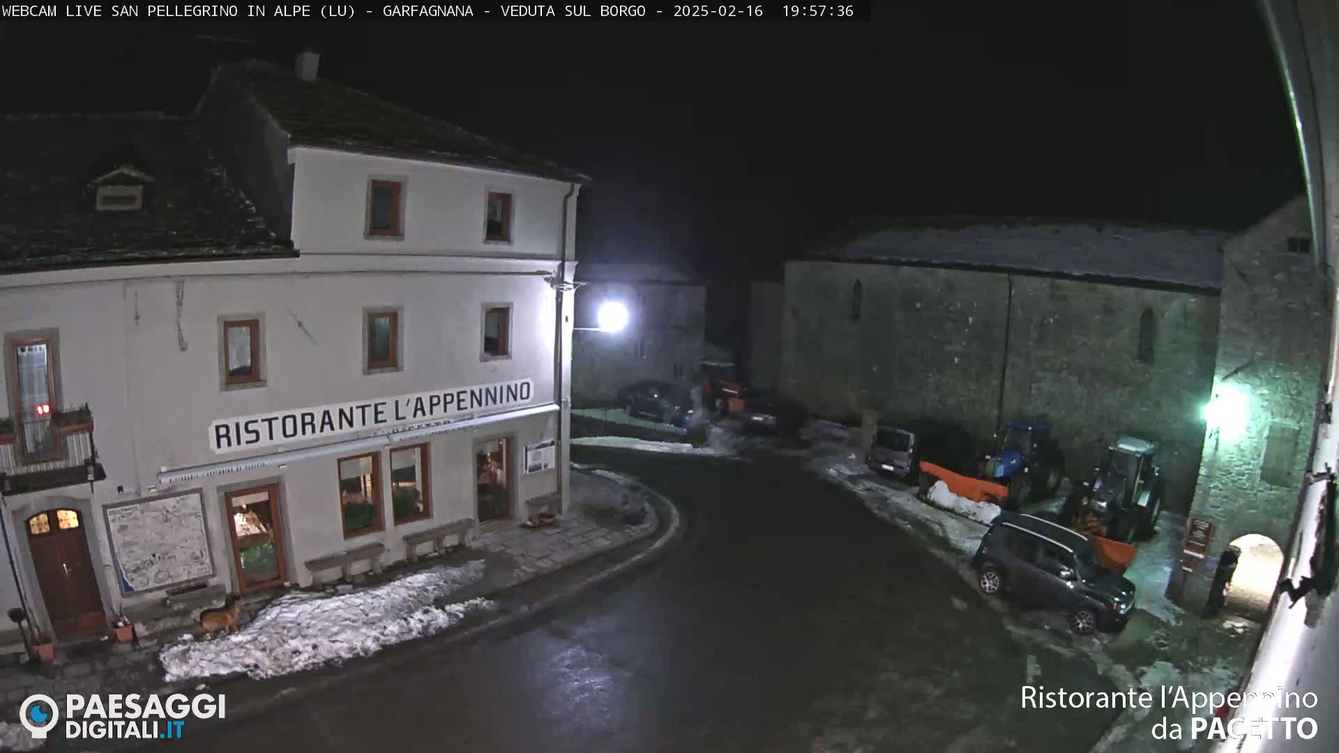 A nighttime view of a snow-dusted town square, featuring a restaurant, parked cars, and tractors, under a dark sky.