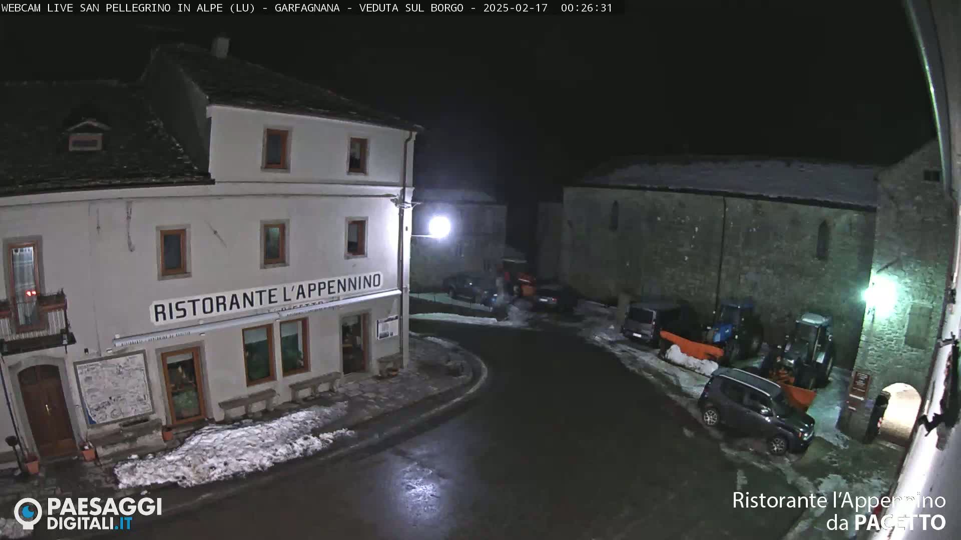 A nighttime view of a village square with snow on the ground, featuring a restaurant and several parked vehicles, under a dark sky.