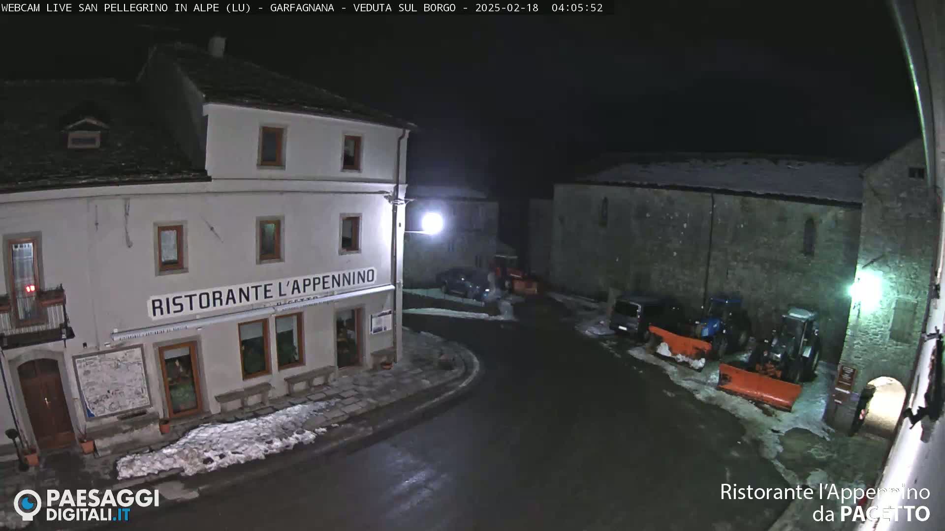 A nighttime, snow-covered village square with a restaurant, parked cars, and snowplows.