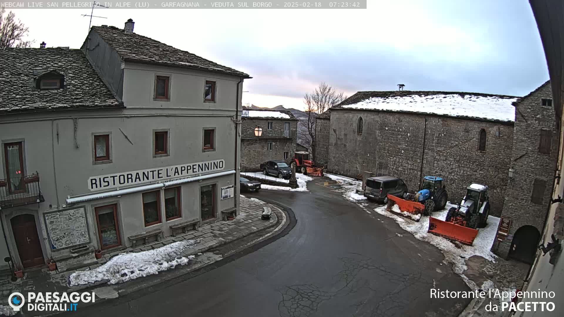 A snow-covered village square with several vehicles, including two tractors with snow plows, parked near stone buildings under a cloudy sky.