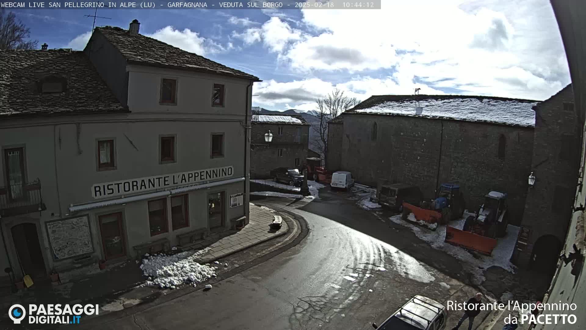 A partly sunny, snowy village square scene shows stone buildings, a restaurant, parked vehicles, and snow-covered patches on the ground.
