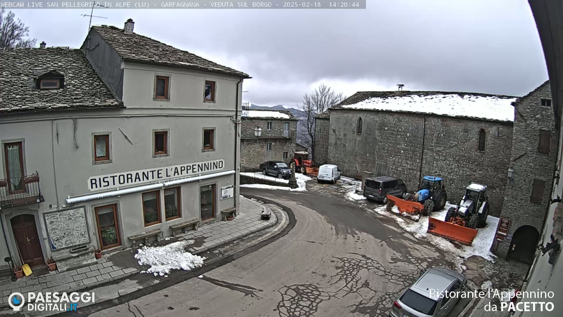 A stone village square with some snow on the ground and parked vehicles, including snow plows, under an overcast sky.