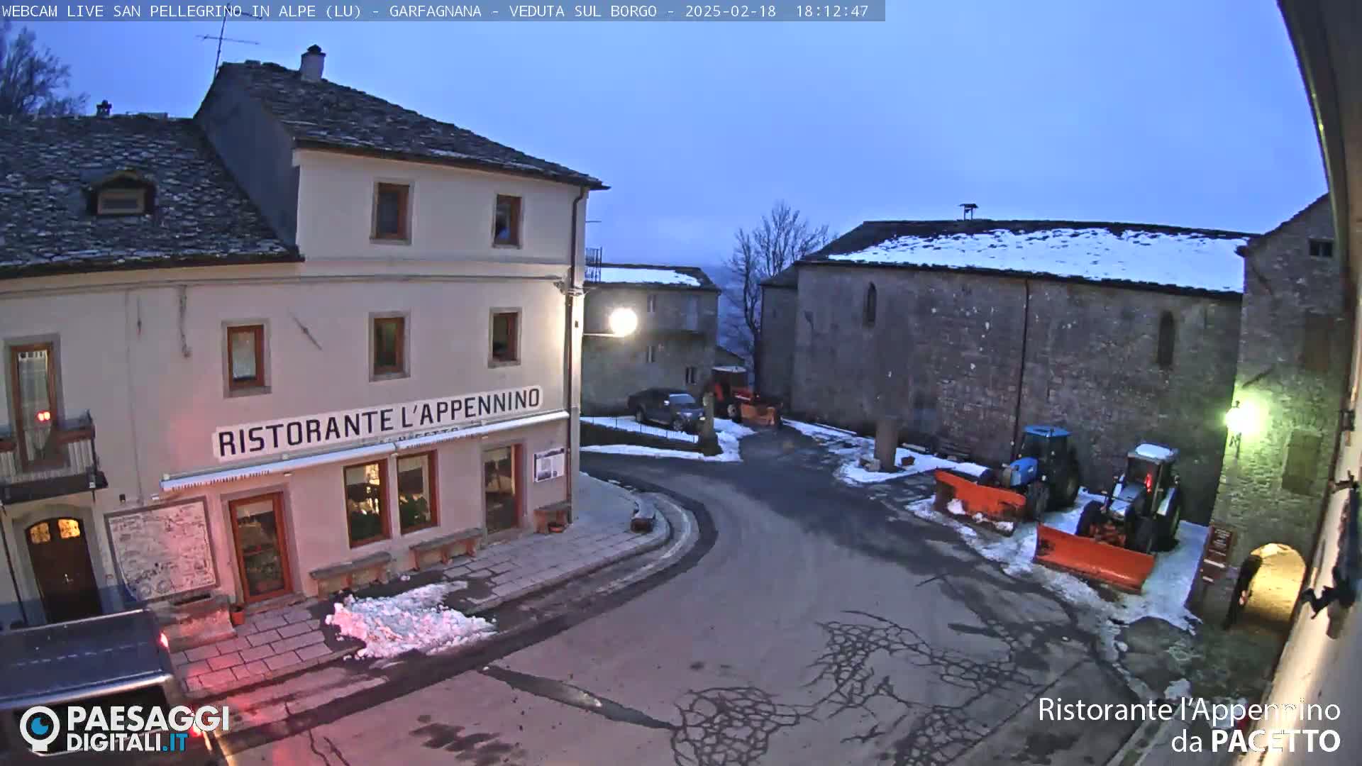 A snow-dusted village square at twilight features a restaurant, several buildings, parked vehicles, and snowplows.