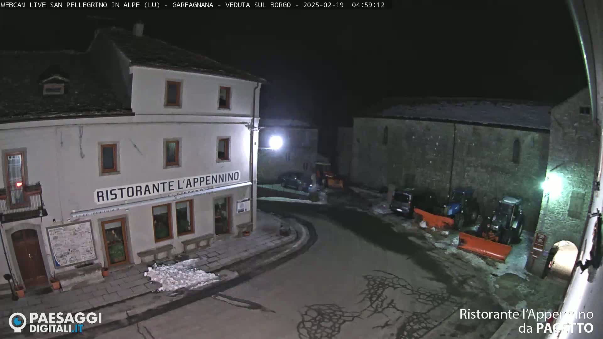 A nighttime view of a snow-dusted town square with a restaurant, several parked vehicles, and snow-clearing equipment.