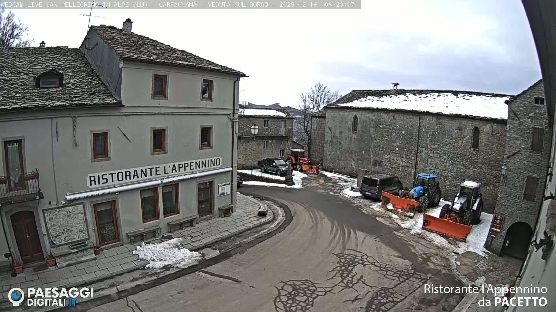A snow-dusted village square with stone buildings, parked vehicles, and snow-clearing equipment under an overcast sky.