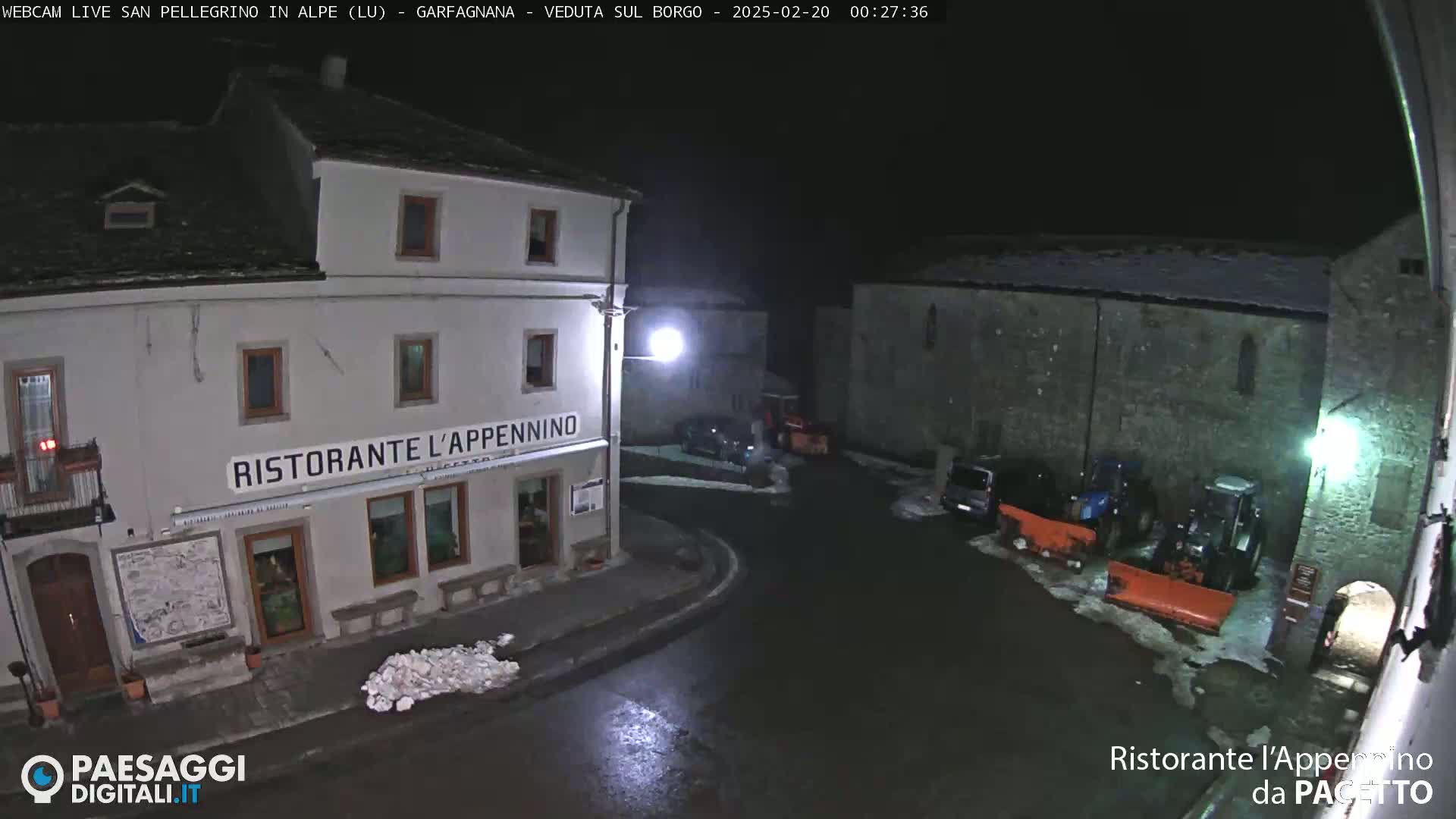 A nighttime view of a snow-dusted town square with a restaurant, several parked vehicles, and snow-removal equipment.