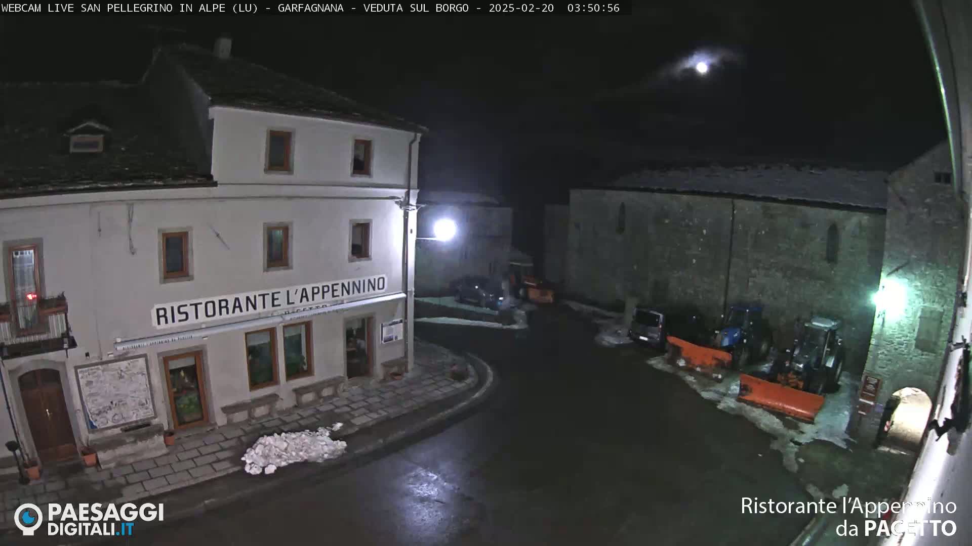 A moonlit nighttime view of a snow-dusted town square with parked vehicles and snow removal equipment near a restaurant.