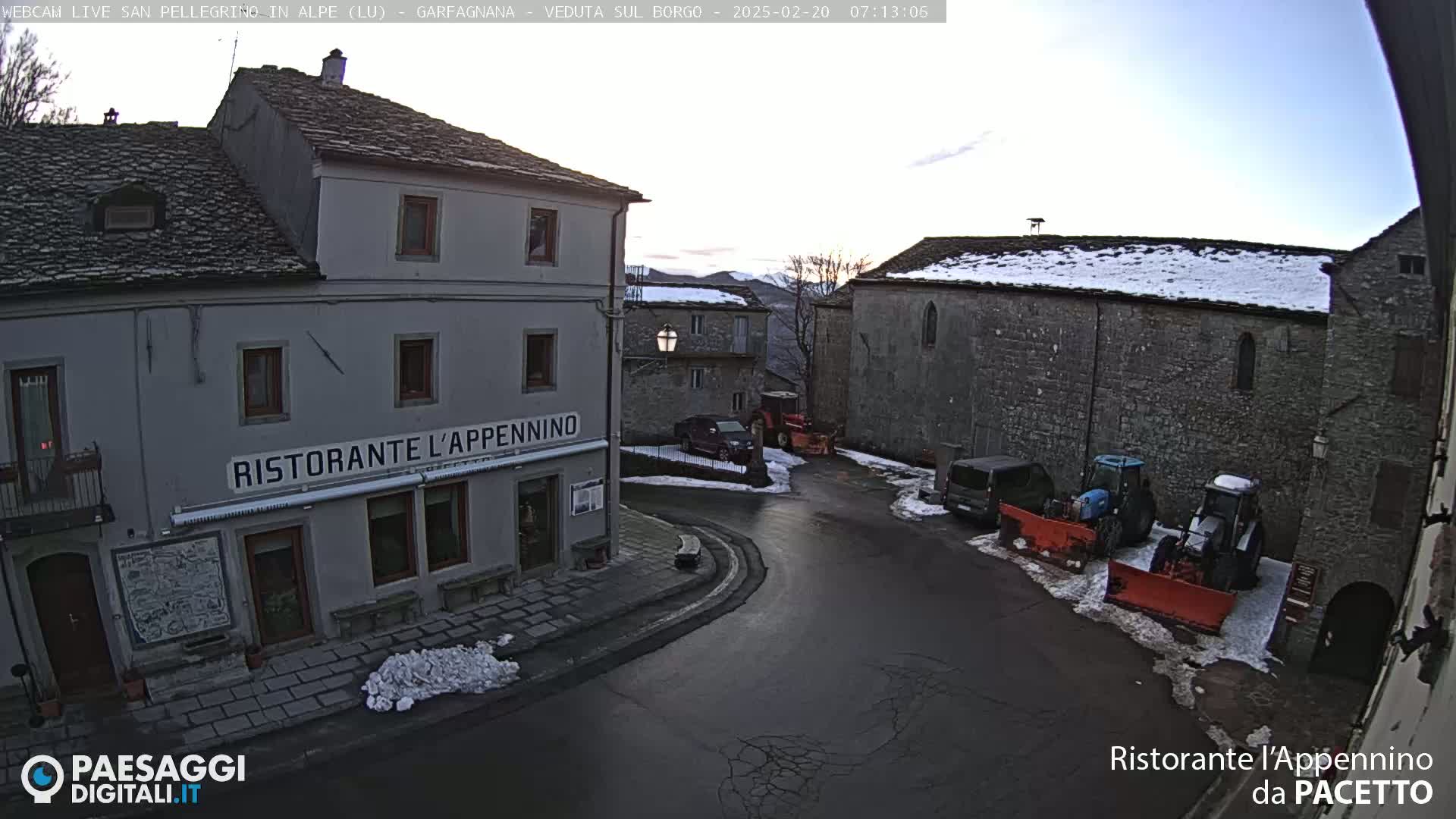 A small town square with snow on the ground and some parked vehicles, including snow plows, is visible under a mostly clear sky.