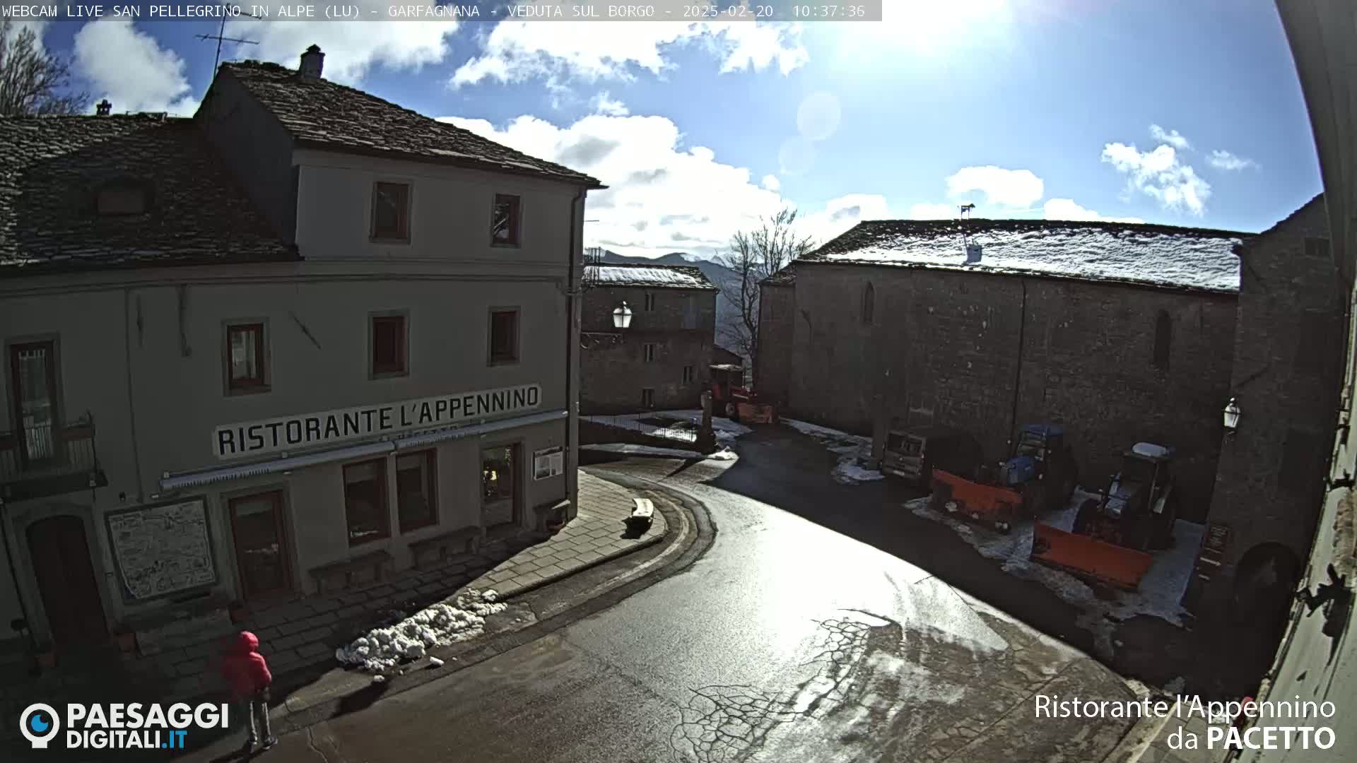 A sunny day reveals a village square with stone buildings, snow on the ground, and some snow removal equipment parked nearby.