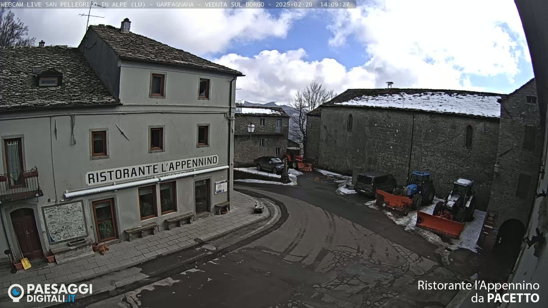 A partly cloudy day reveals a small town square with stone buildings, parked vehicles, and snow on the ground and some rooftops.