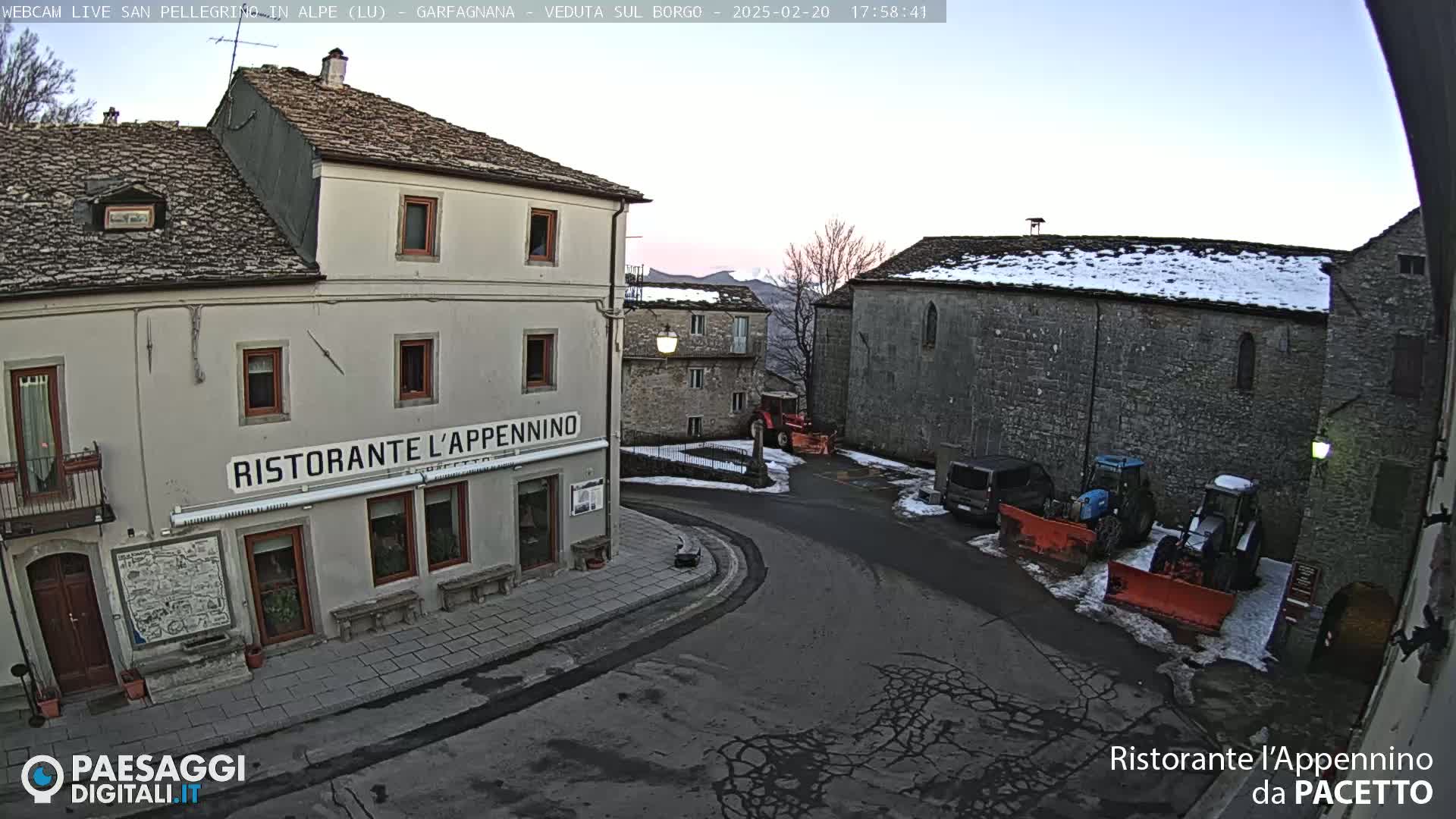 A village square with stone buildings, some snow on the ground, and several tractors and vehicles parked nearby under a twilight sky.
