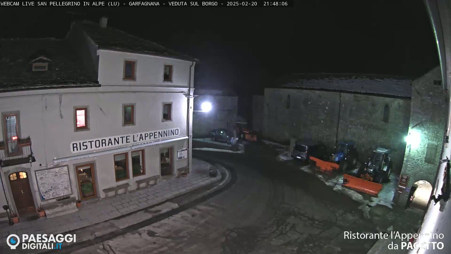 A snow-covered nighttime town square features a restaurant, parked vehicles, and snow-clearing equipment.