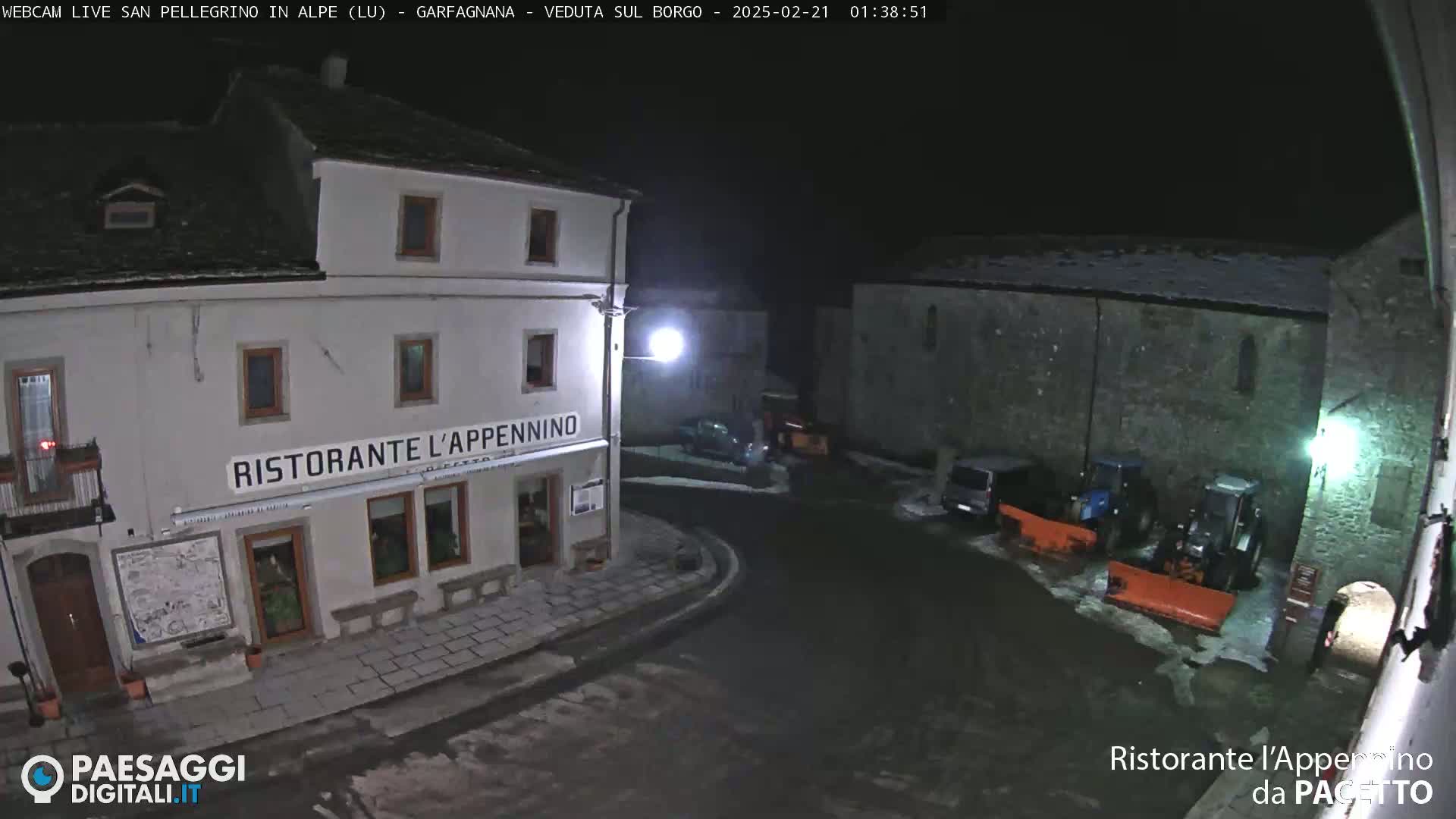 A nighttime, snow-dusted village square features a restaurant and parked vehicles, with tractors and snowplows near a stone building.