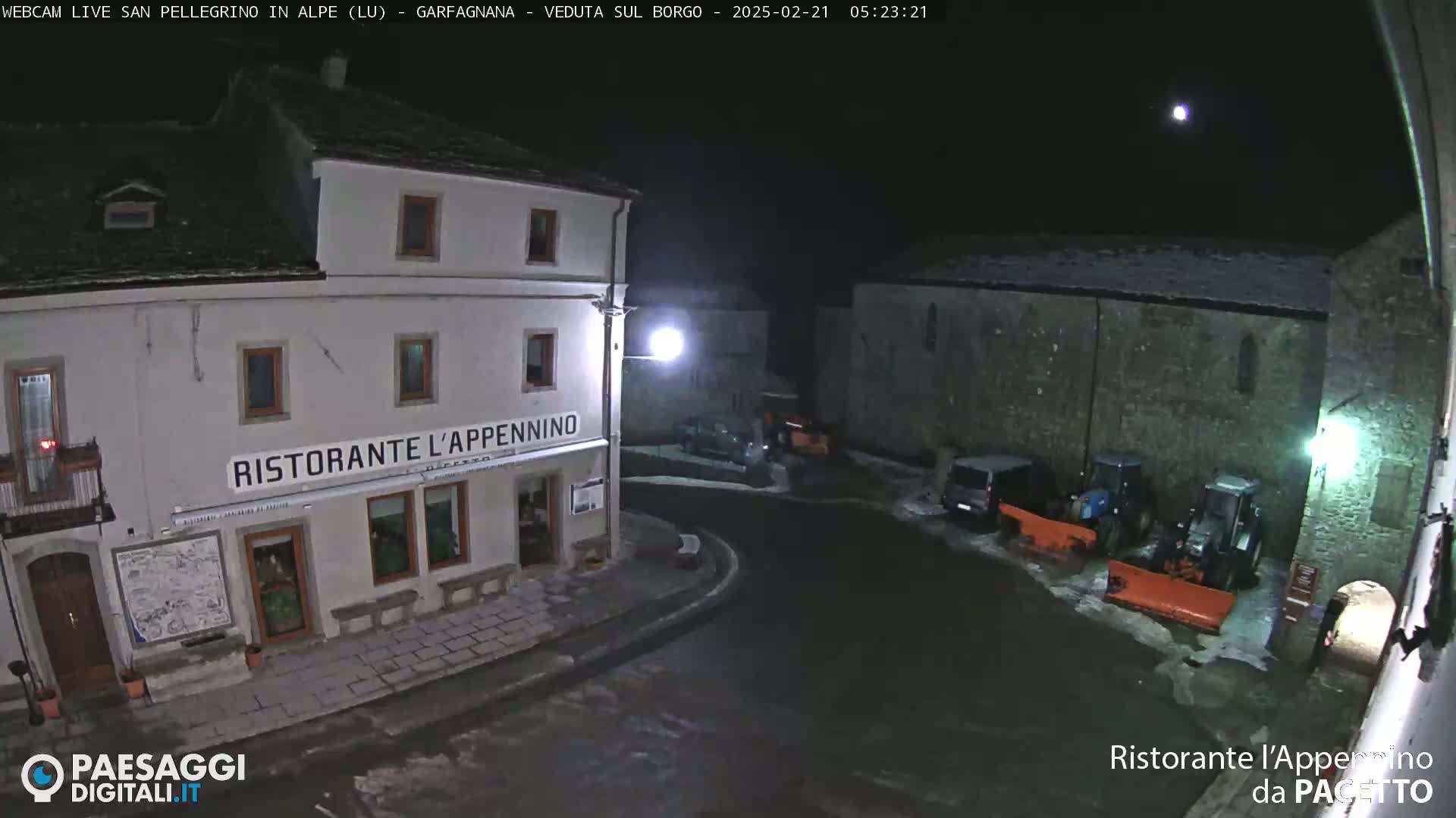 A nighttime view of a snow-covered town square with a restaurant, several vehicles, and snowplows.