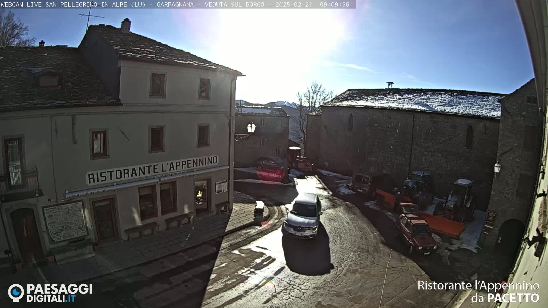 A sunny, snow-dusted village square features several buildings, some vehicles, and a restaurant.