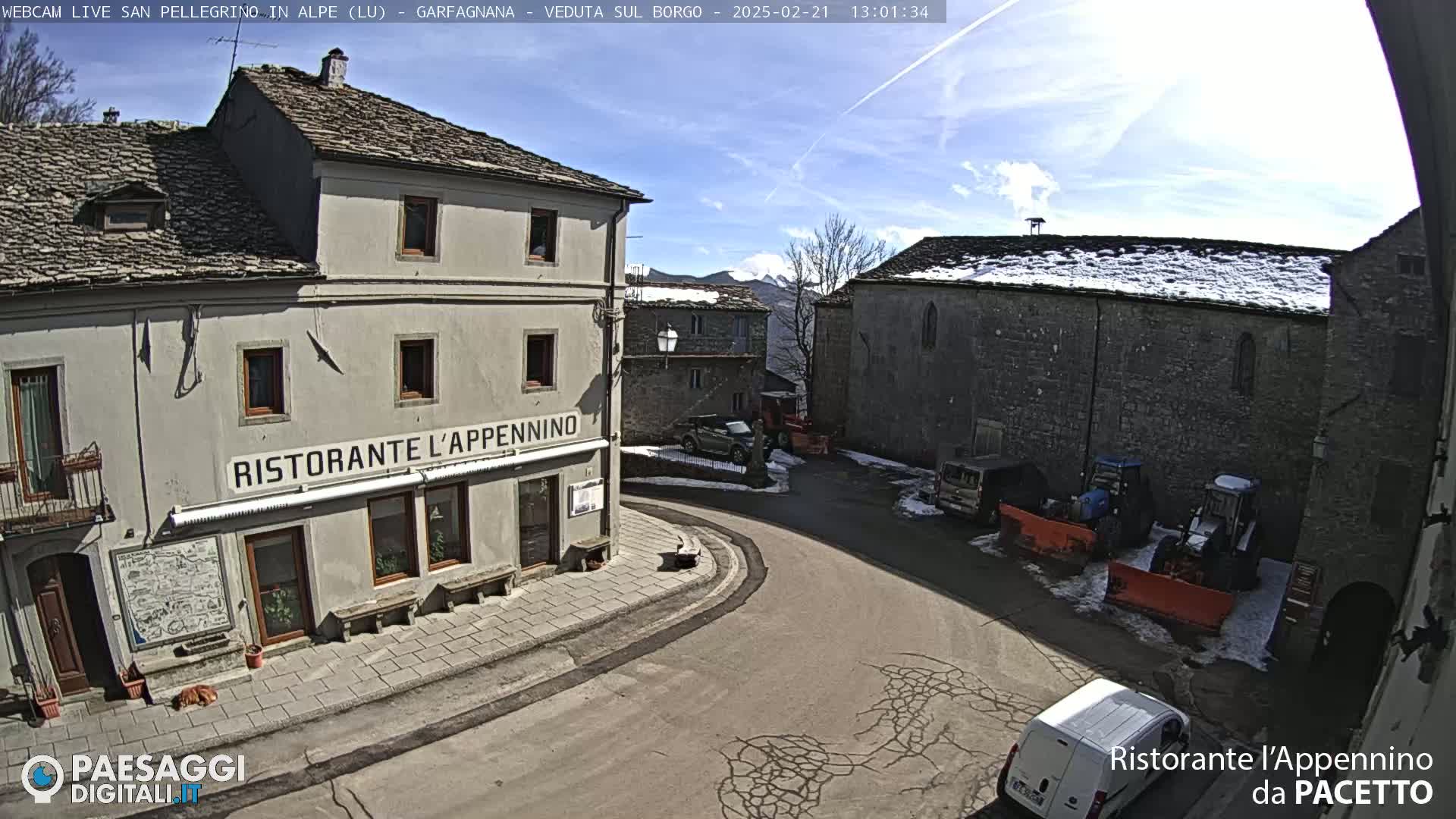 A sunny village square with stone buildings, some snow on the ground, and several vehicles parked nearby.