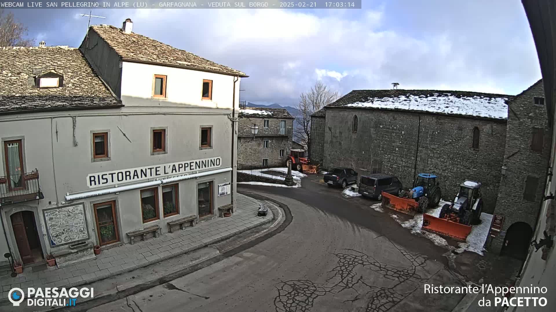 A partly cloudy day reveals a village square with stone buildings, some snow on the ground and rooftops, parked vehicles, and tractors with snowplows.