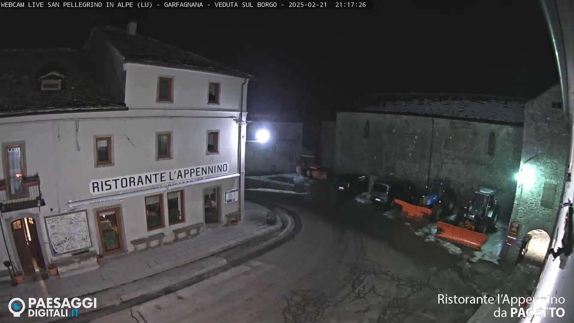 A nighttime view of a town square with a restaurant, several parked cars, and snow-clearing equipment.