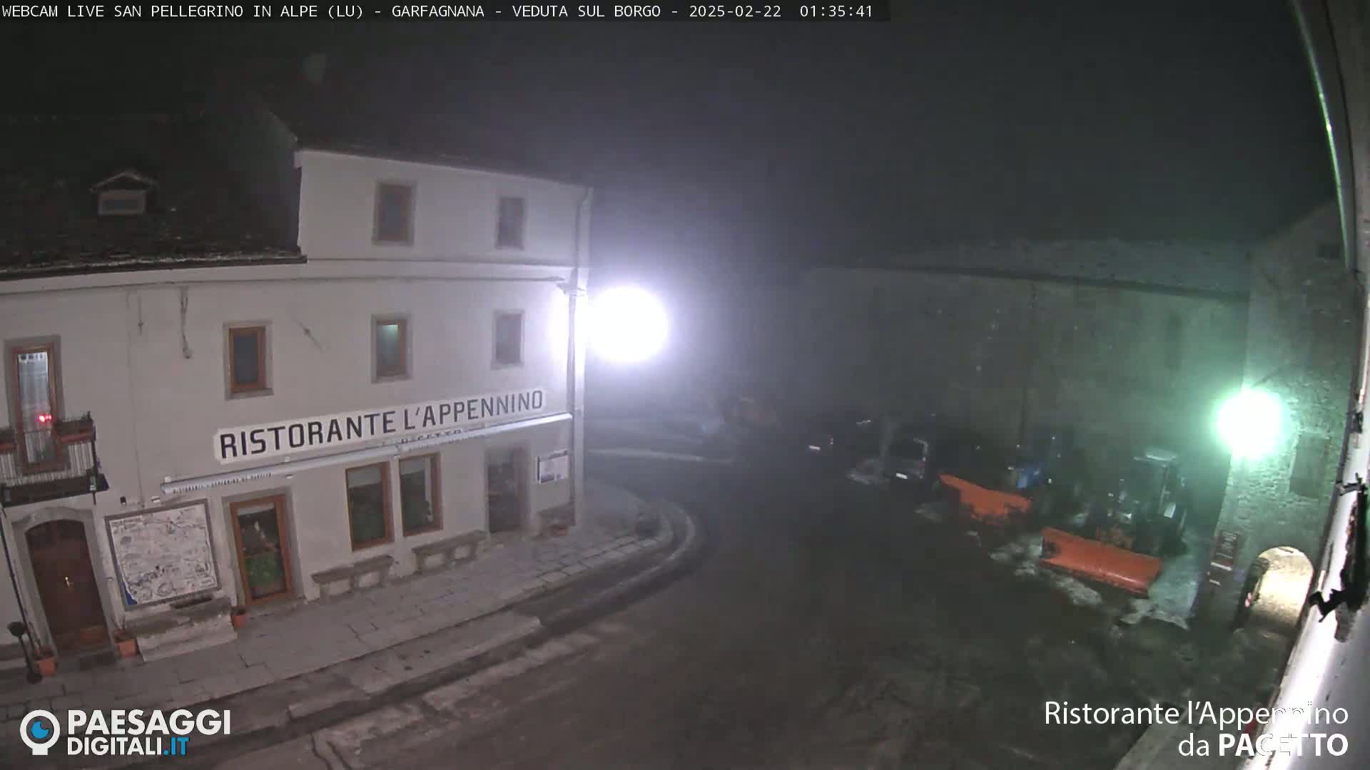 A nighttime, foggy view of a town square with a restaurant, snow plows, and parked vehicles.