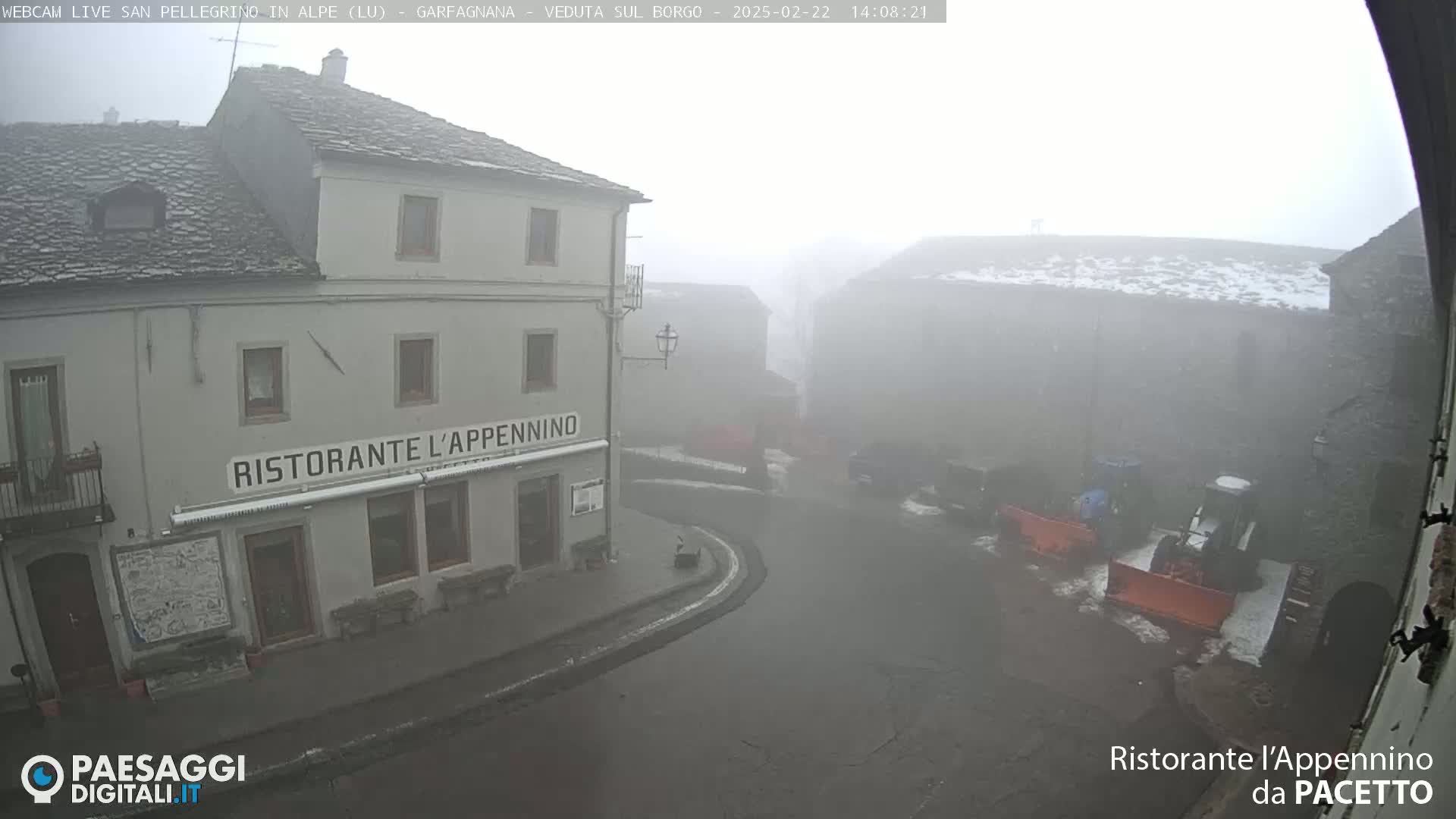 A foggy village square is seen, with a restaurant and snow-covered buildings in the background, and snow plows parked in the street.