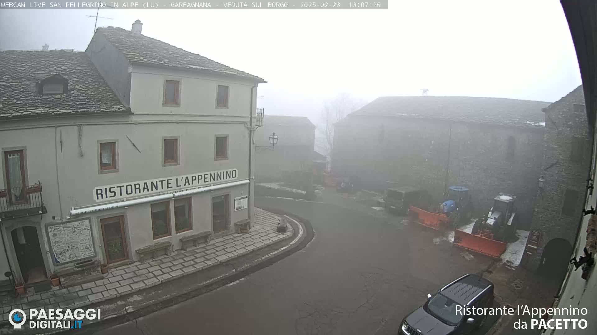 A foggy village square with stone buildings, a restaurant, and snowplows is seen from a high vantage point.