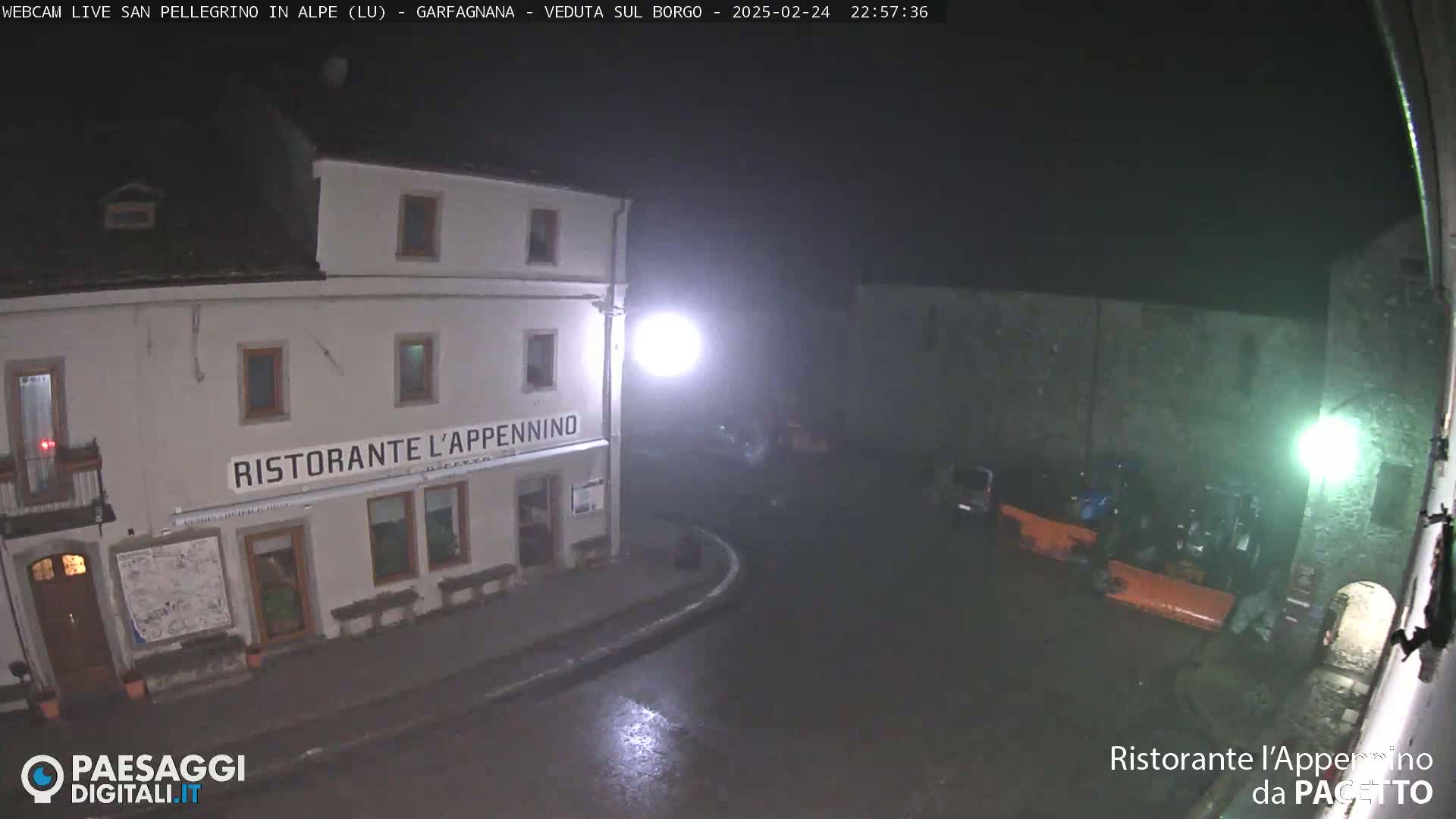 A nighttime view of a town square with a restaurant, some parked vehicles, and wet pavement under dim lighting.