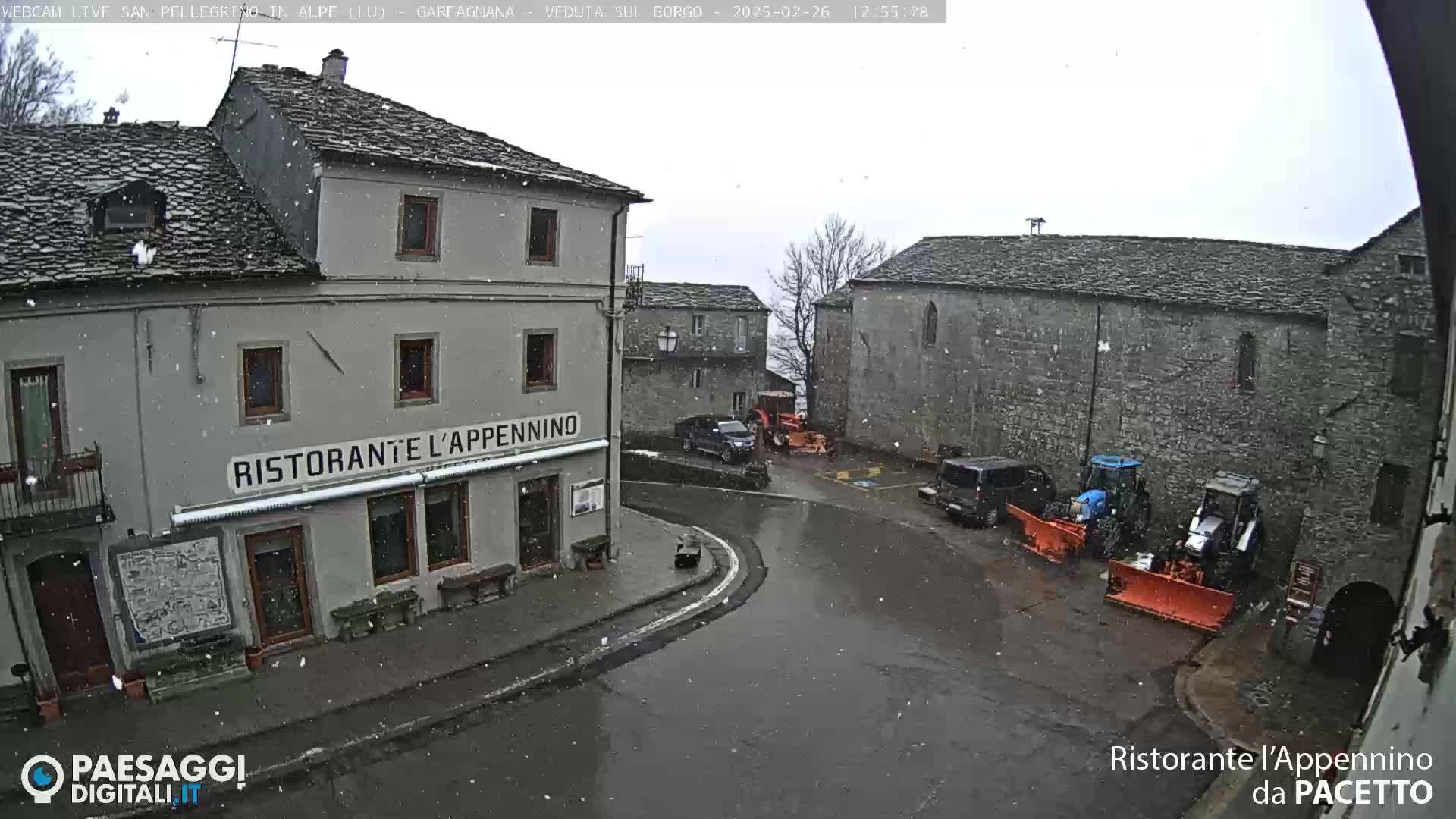 A snow-covered village square with stone buildings, a restaurant, several vehicles, and two snow plows.