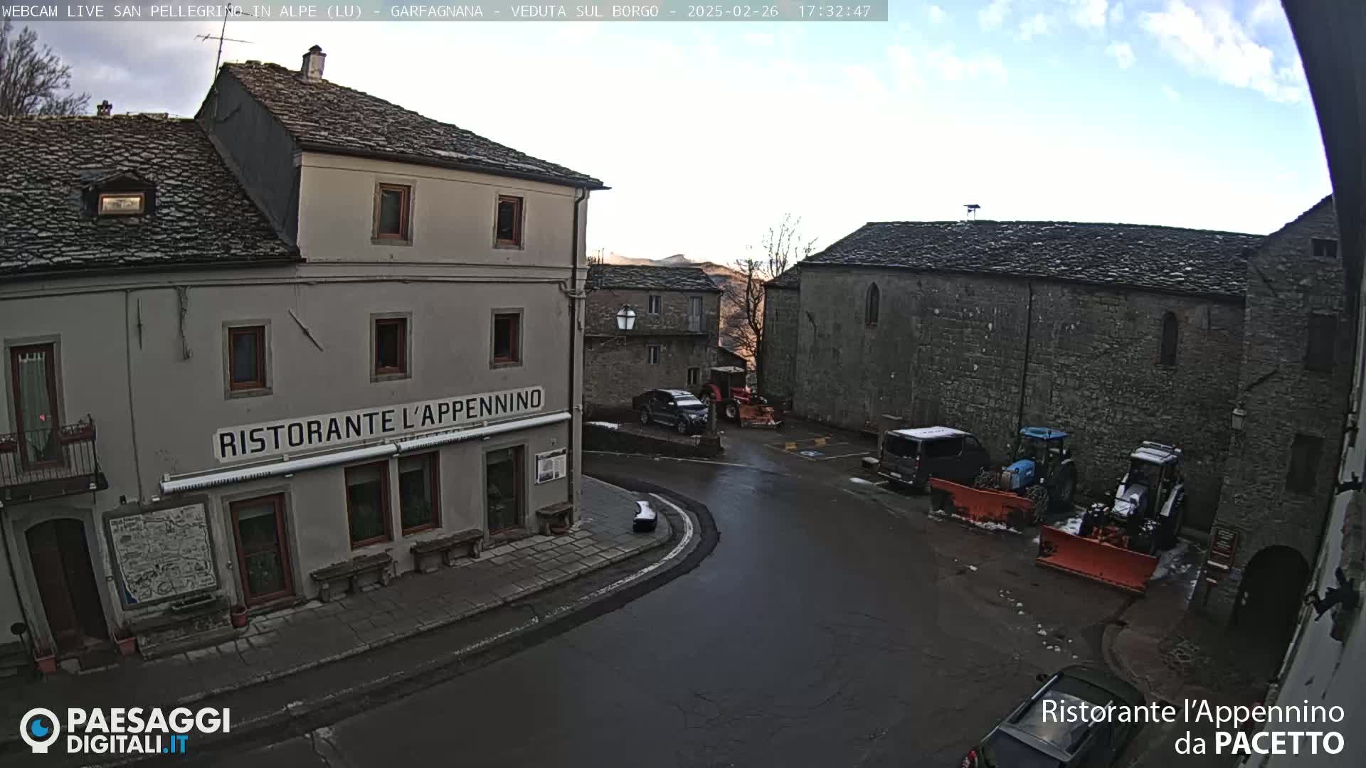 A partly cloudy daytime view of a town square with several stone buildings, some vehicles, and farm equipment, including tractors with snow plows.