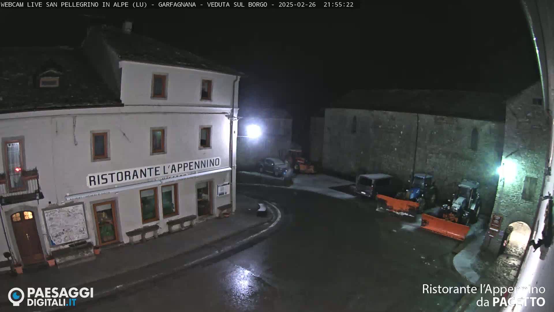 A nighttime view of a town square with snow-covered ground, featuring a restaurant, several parked vehicles, and snowplows.