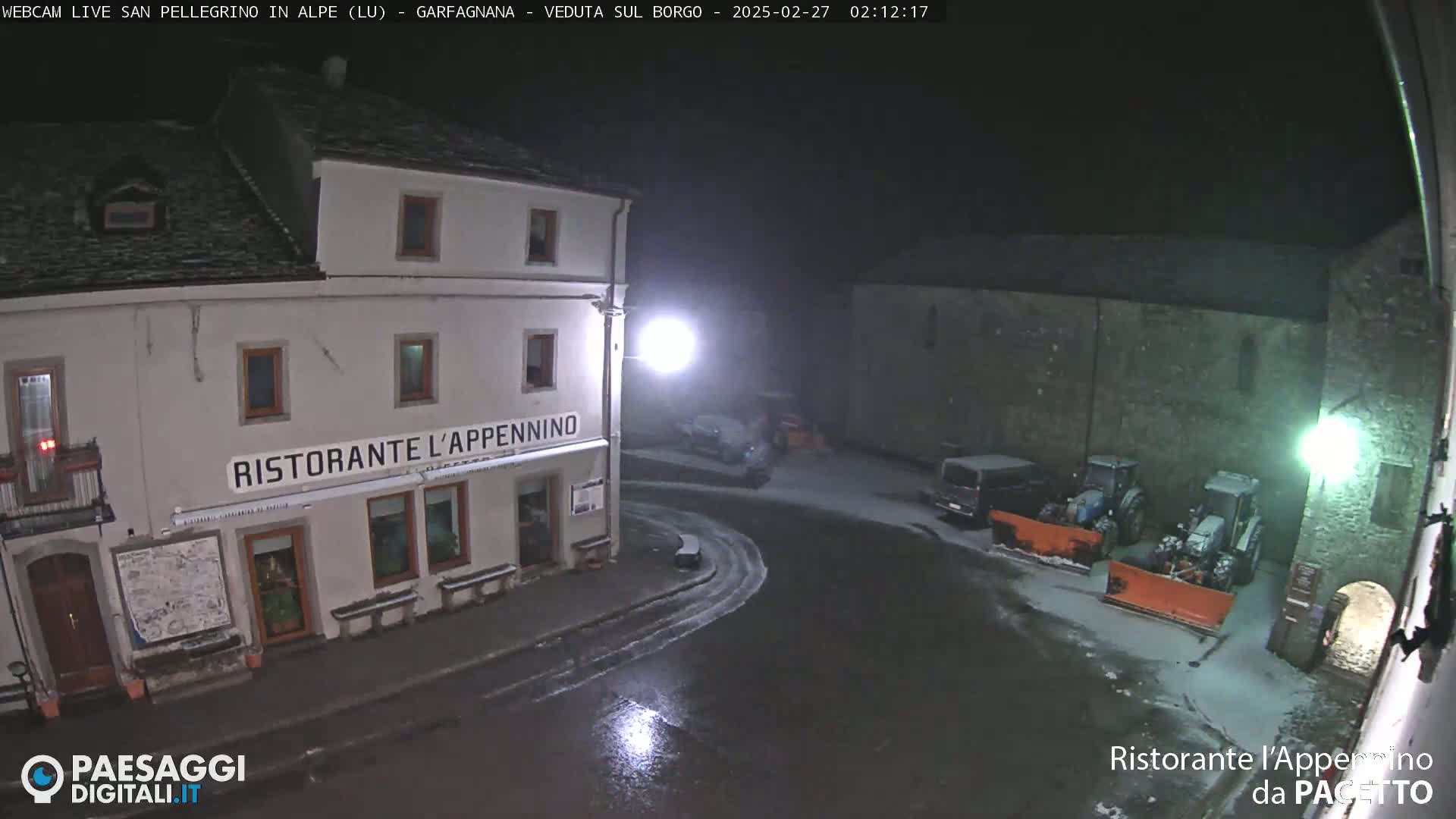 A snow-covered town square at night, illuminated by streetlights, shows a restaurant and several parked vehicles, including snow plows.
