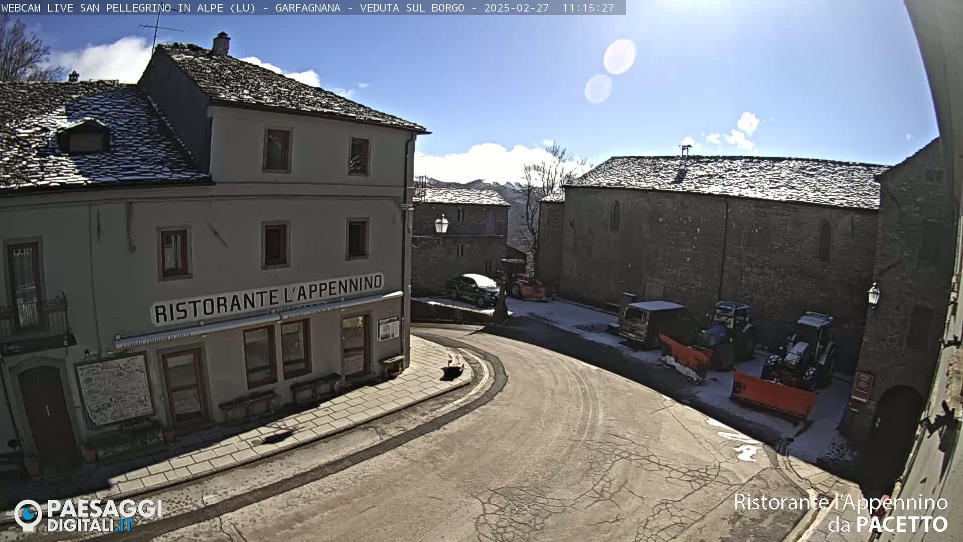 A sunny, snow-dusted village square features stone buildings, a restaurant, parked vehicles, and snow-clearing equipment.