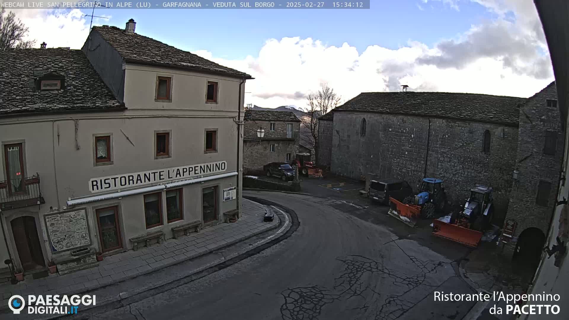 A partly cloudy day reveals a quaint village square with stone buildings, a restaurant, and snow removal equipment.