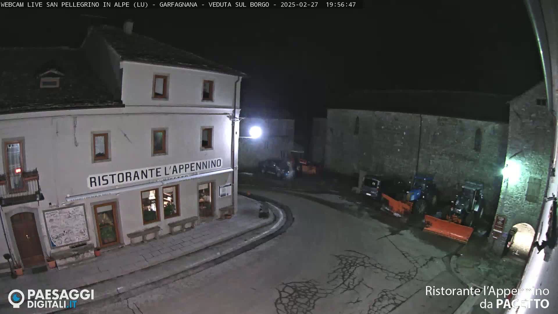 A nighttime view of a town square with a restaurant, several parked vehicles, and snowplows.