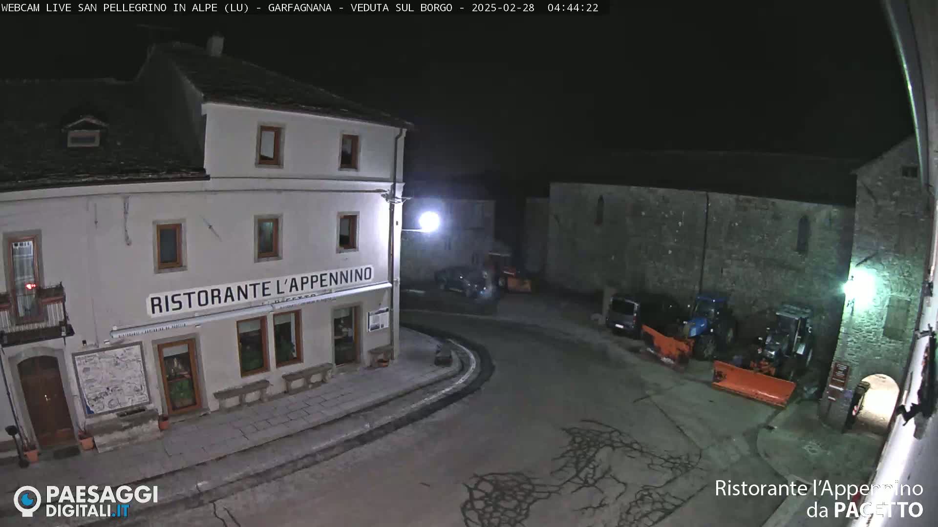 A nighttime view of a town square with a restaurant, some parked vehicles, and snowplows.