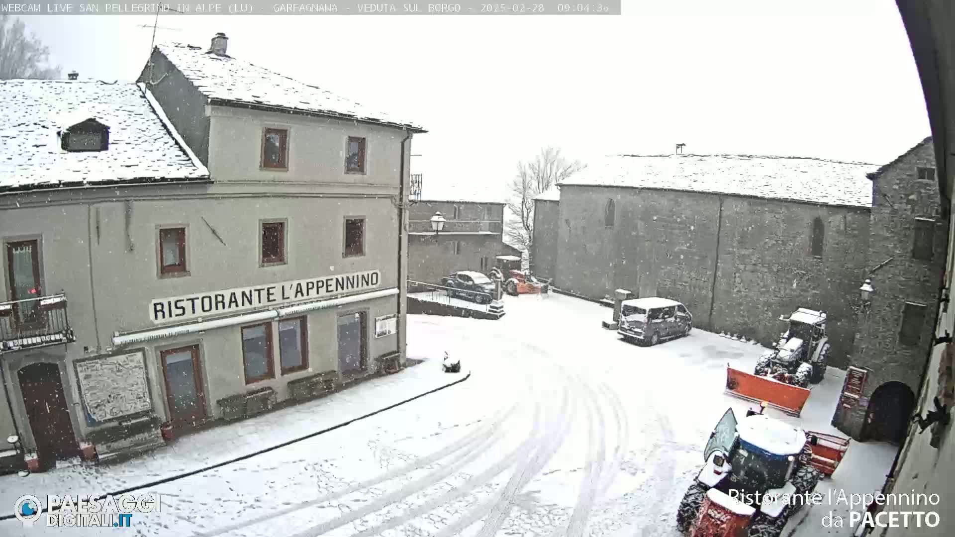 A snow-covered village square with several buildings, a few parked cars, and two tractors, one of which is a snowplow, during a light snowfall.