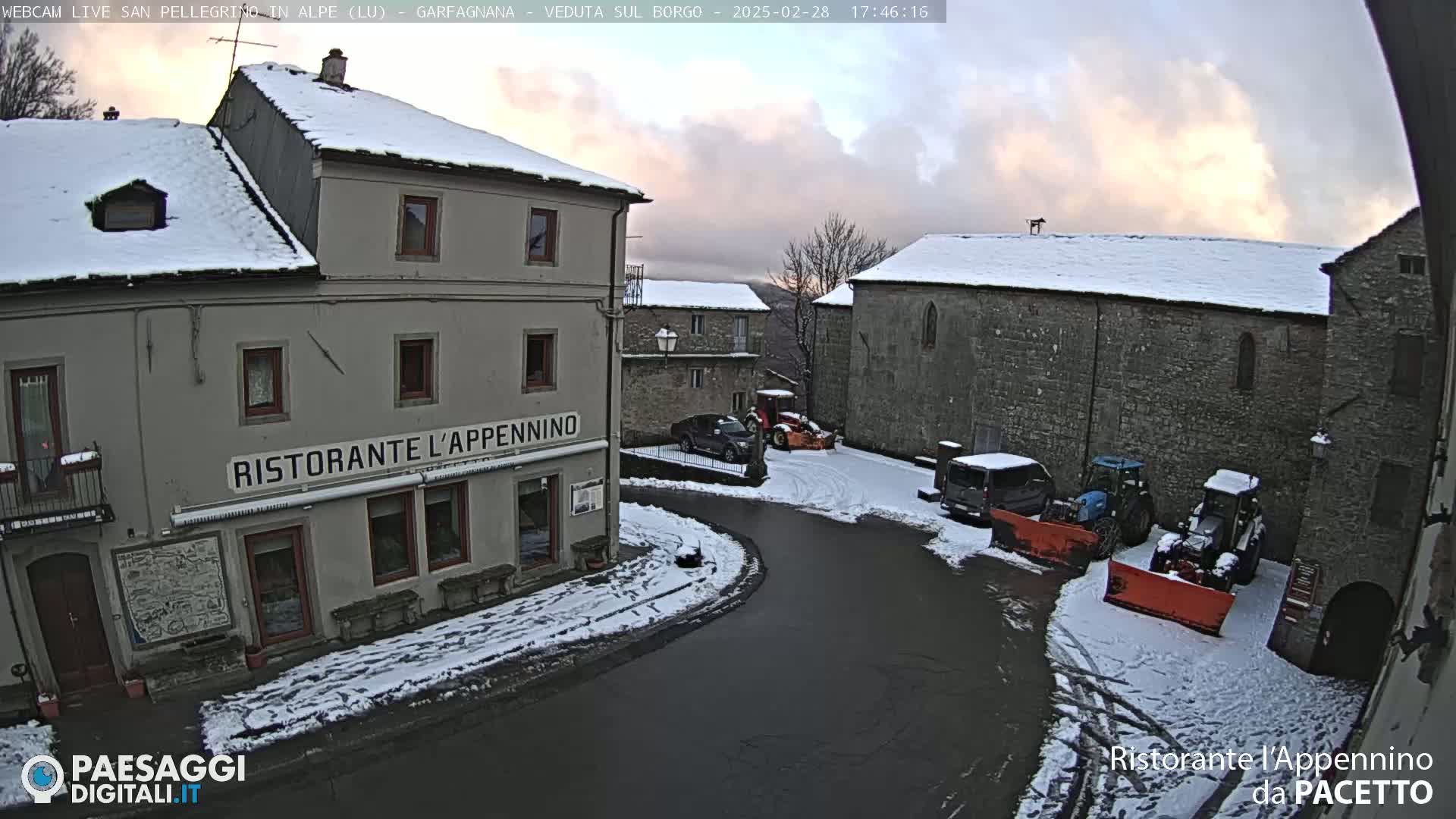A snow-covered village square with several buildings, a few vehicles, and snow removal equipment under a partly cloudy twilight sky.