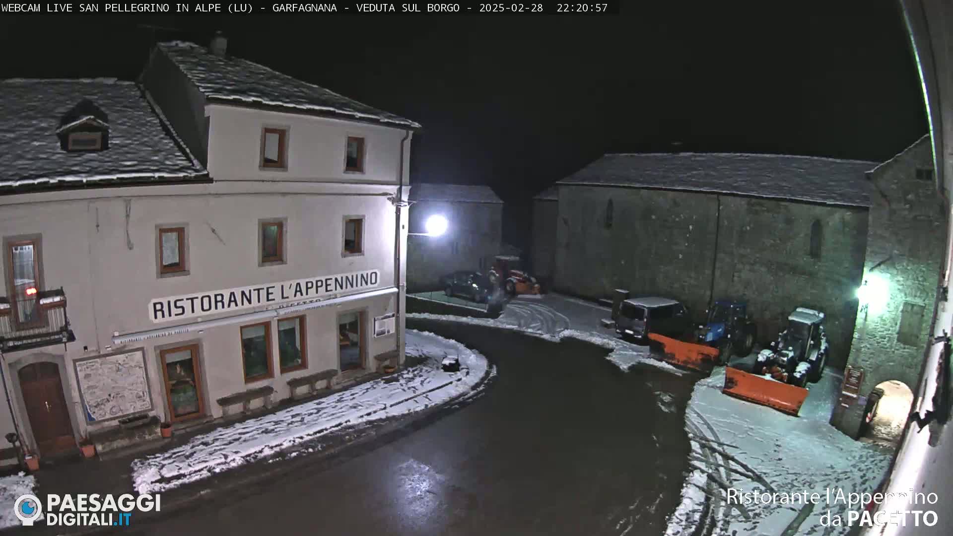 A snow-covered village square at night is illuminated, showing a restaurant, some parked vehicles, and snow removal equipment.