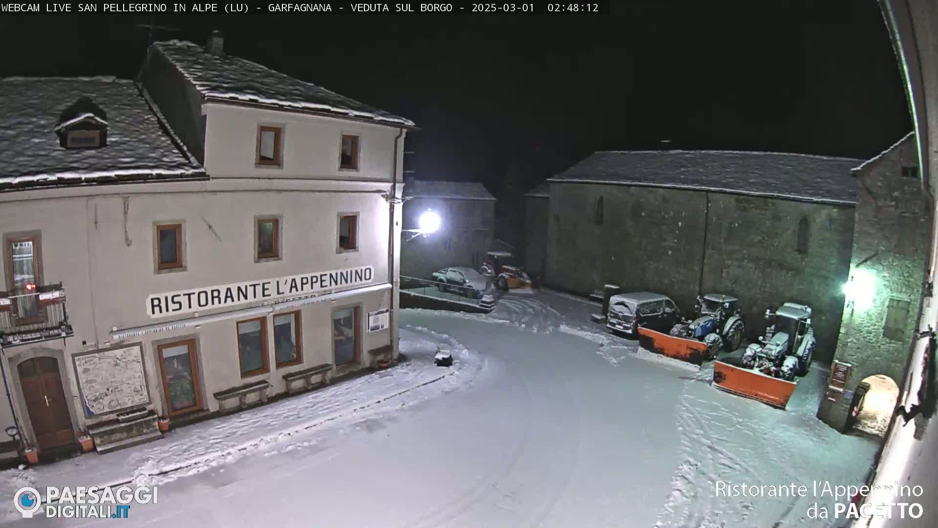 A snow-covered town square at night, with a restaurant, parked vehicles, and snow-clearing equipment.