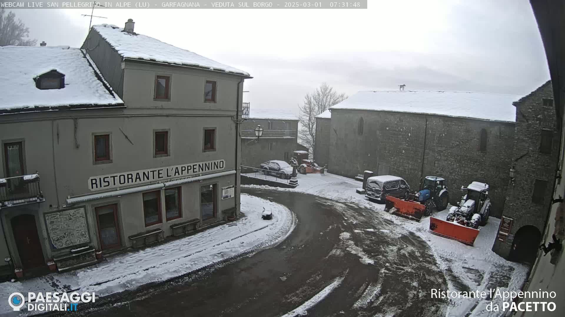 A snow-covered village square with several buildings, parked cars, and snow plows.