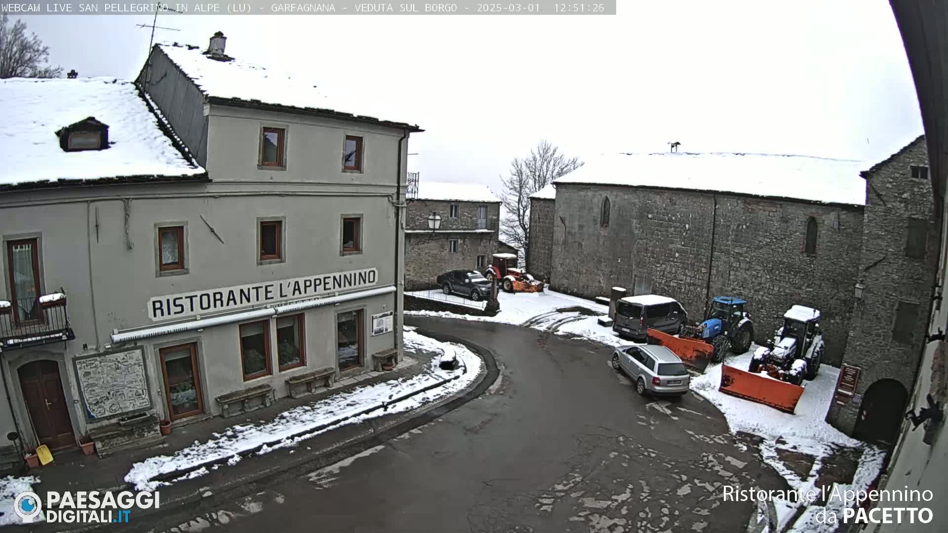 A snow-covered village square with several cars and snow removal equipment parked around a restaurant.