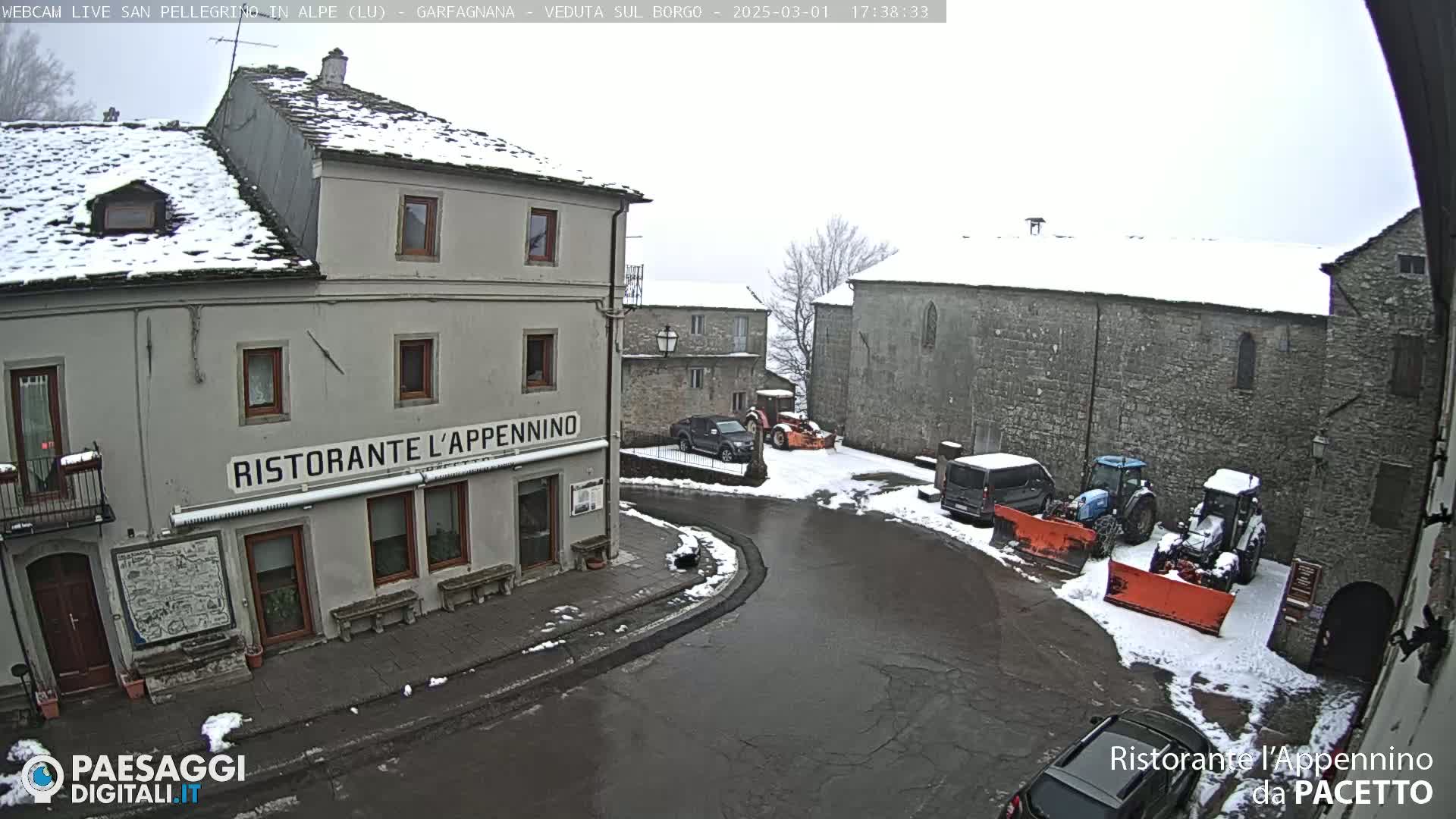 A snow-covered village square with several vehicles, including tractors and a van, parked near stone buildings, one of which is a restaurant.